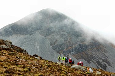 Picture of walkers in front of a steep mountain, clouds lifting on the mountain