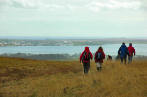 Picture of walkers coming over a hill, a sea loch coming into view with a village on the other side