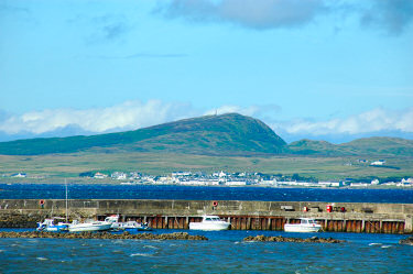 Picture of a view over sea loch behind a pier, a coastal village under a hill visible on the other side